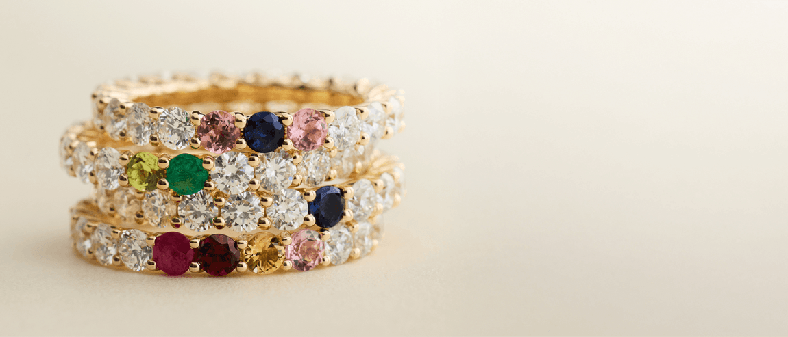 Stack of gold rings with colorful gemstones arranged on a neutral background.